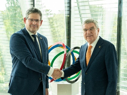 Adam Kosa and Thomas Back share a friendly handshake in front of a table adorned with an Olympic symbol, with a scenic view through the window.